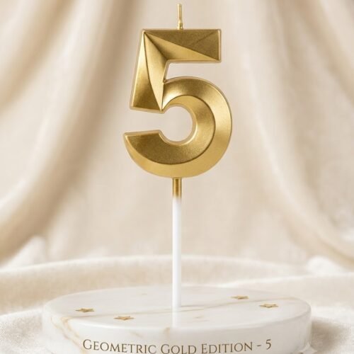A close-up photograph of a geometric, faceted gold number "5" birthday candle standing upright on a round, polished white marble plinth. A small gold star is placed next to the candle's base. The background is a soft, blurred, cream-colored fabric draped behind the display. The image uses elegant, high-quality studio lighting. The marble base has the text 'GEOMETRIC GOLD EDITION - 5