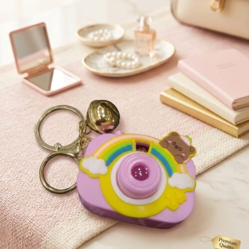 cute pastel purple camera shaped keychain featuring a rainbow design, small brown bear character, and a golden jingle bell on a pink textured runner