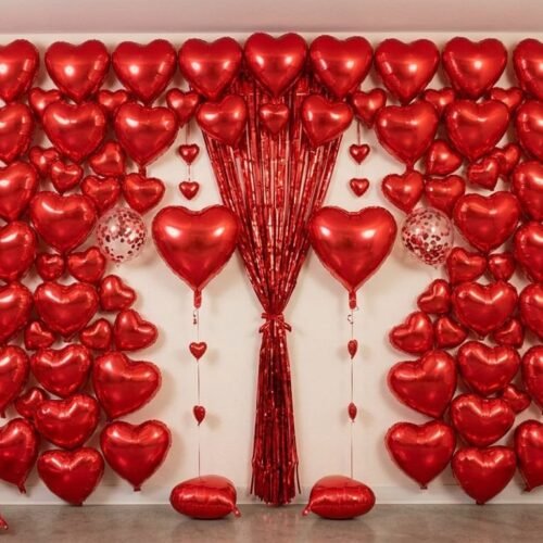romantic valentine decoration setup featuring multiple red heart foil balloons, metallic fringe curtains, and confetti balloons against a white wall